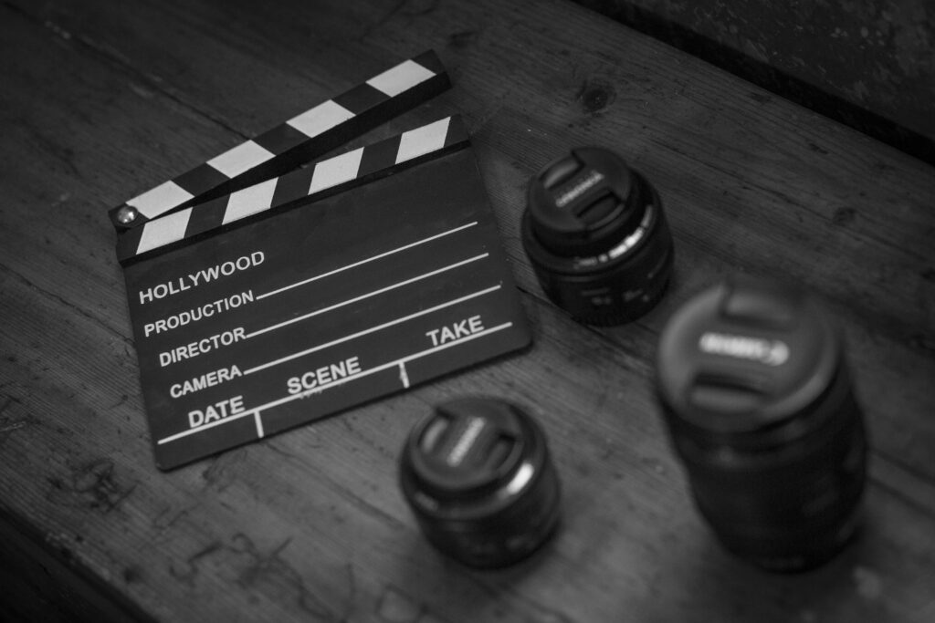 pexels photo 5627838 5627838 Monochrome image featuring a clapperboard and camera lenses on a wooden table.